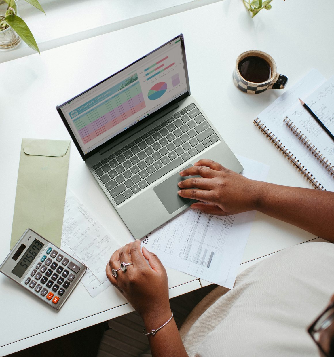 Person working on financial spreadsheets on a laptop at a desk with a notebook, calculator, and coffee cup.